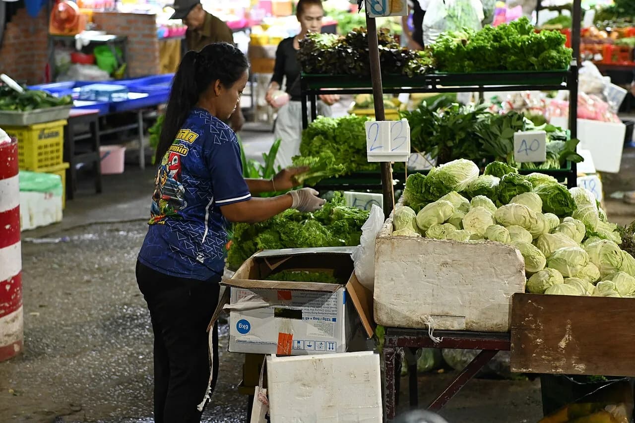 DFC 1793 A vendor arranges fresh greens and cabbages at an open-air market stall while shoppers browse in the background