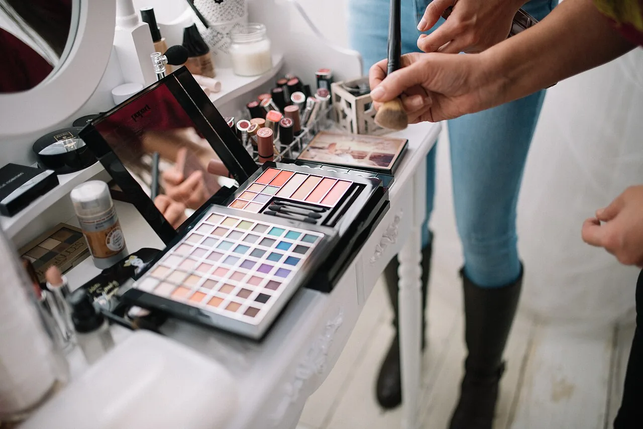 Hands of a makeup artist are shown holding a brush while colorful makeup palettes are arranged neatly on a clean table i
